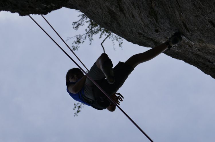 A climber hanging upside down from the underside of an overhanging rock arch, silhouetted against the sky.