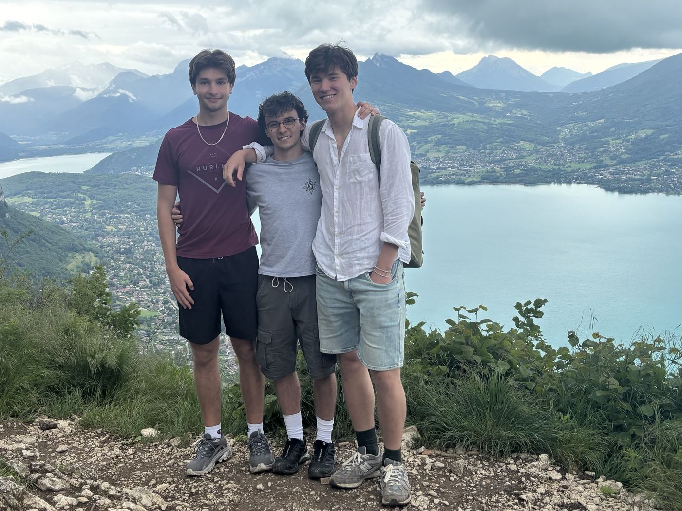 Three friends standing on a rocky lookout above Lake Annecy, with turquoise water, mountains, and heavy clouds behind them.