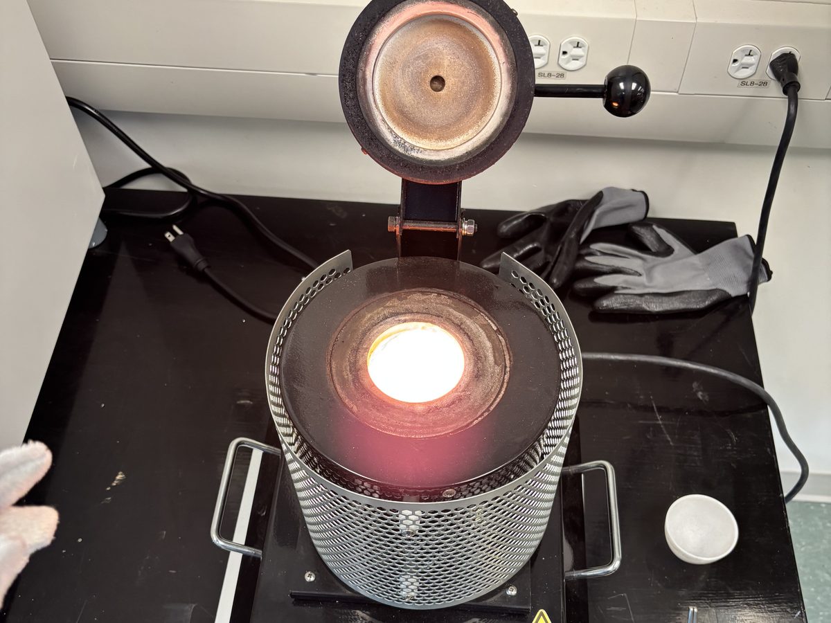 A benchtop melting furnace in a lab with its lid swung open, the crucible inside glowing orange, with tongs and heat-resistant gloves beside it.