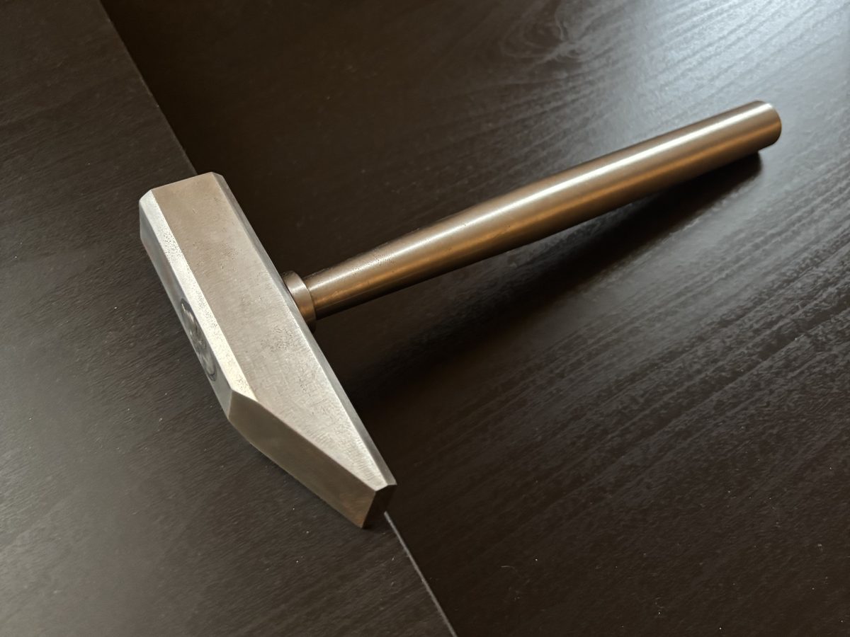 A steel hammer with a faceted head and a turned round handle, lying on a dark wood desk.
