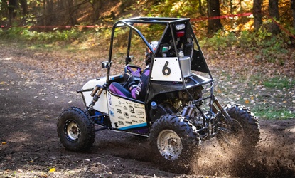Western Baja SAE's car number 6 sliding through a dirt corner on a wooded course, kicking up mud, with the driver in a purple suit.