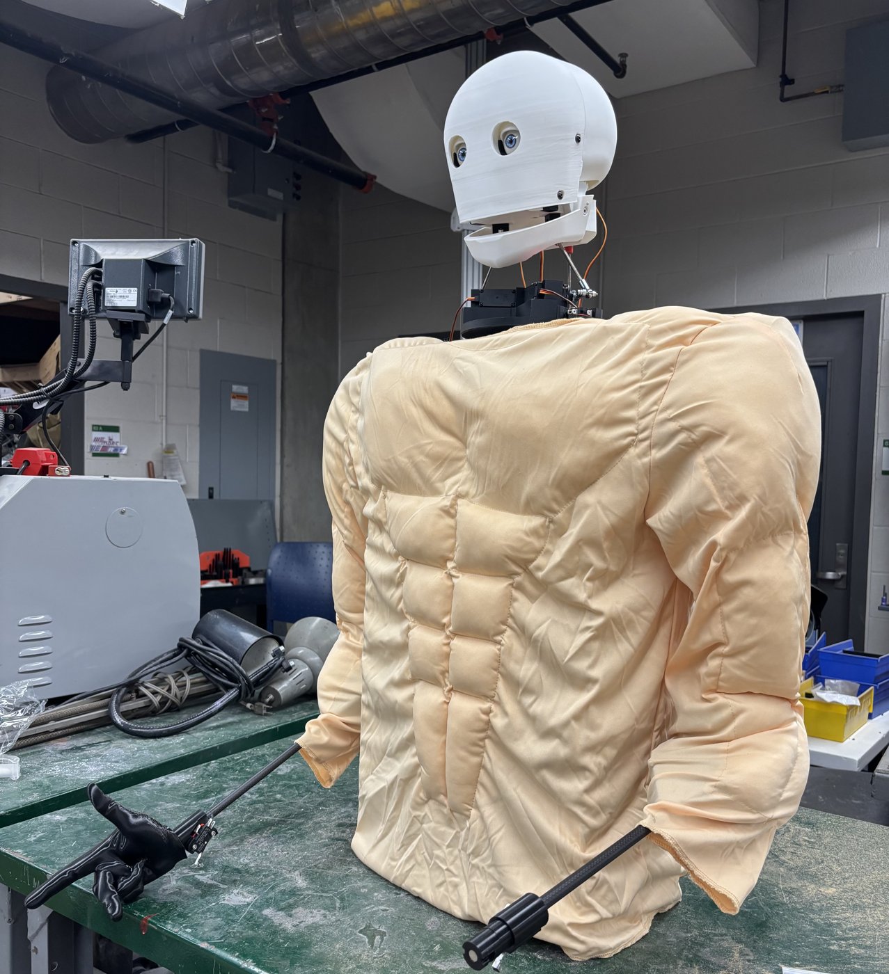 Baker Bot from behind, wearing a padded beige muscle suit over the torso and arms, waist-up on a workbench.