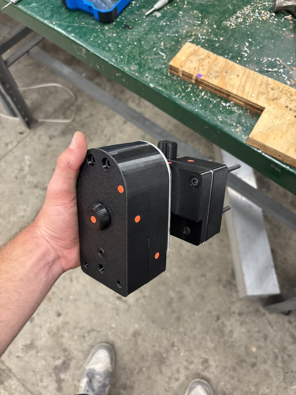 3D-printed shoulder housing held in hand, with a knurled indexing plunger knob and the edge of the aluminum indexing plate visible.
