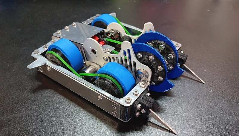 A finished 1 lb vertical spinner combat robot on a workbench: an aluminum chassis with four blue wheels driven by green belts, twin blue weapon discs at the front, and angled wedgelets under the discs.
