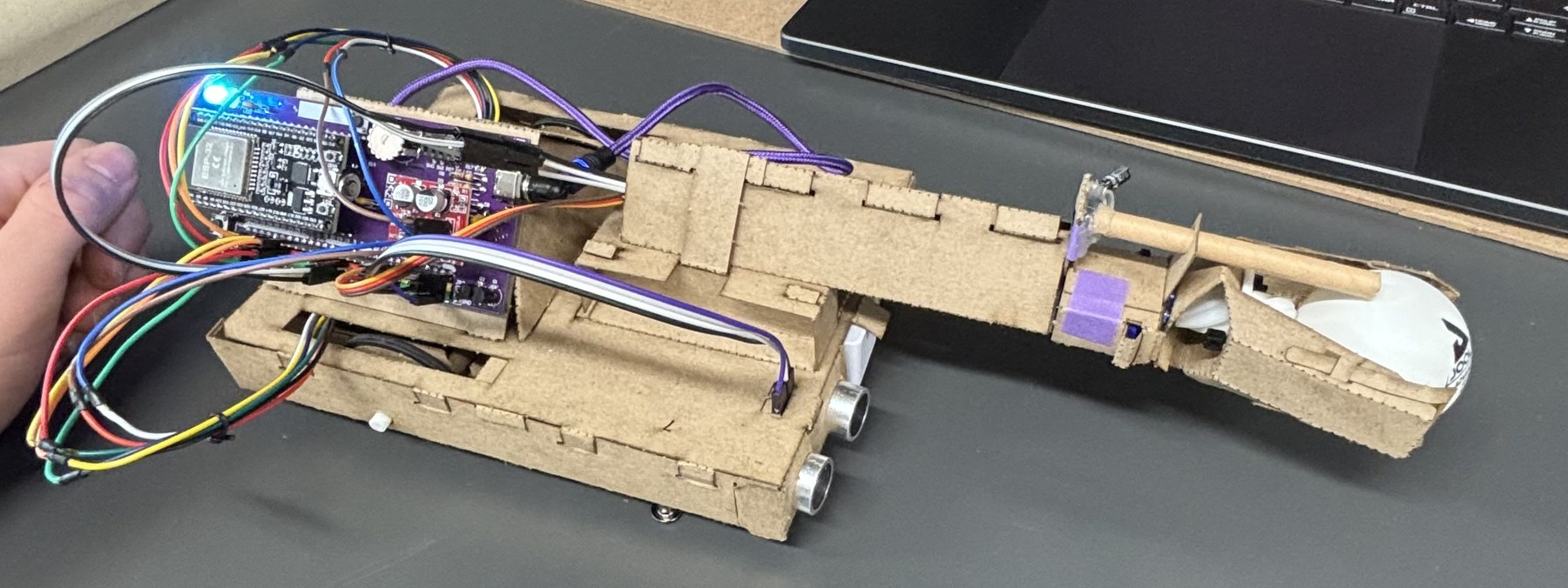 The finished robot on a desk: a laser-cut hardboard chassis with an ESP32 control board and wiring on top, two ultrasonic sensor eyes at the front, and a long arm ending in a claw.