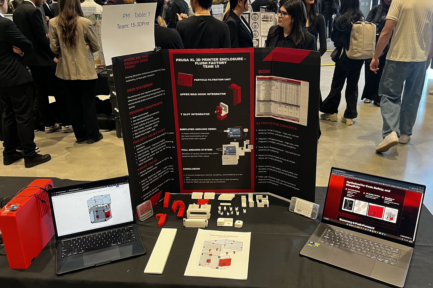 The team's display table at the design competition: a black and red poster board behind a row of 3D printed enclosure parts, a box of magnets, a box of M3 screws, and a printed copy of the build manual.